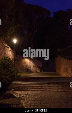 Tranquilla strada acciottolata con lampione luminoso e mura di pietra medievali durante l'ora blu della sera nella storica Roma, Italia Foto Stock