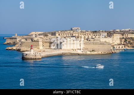 Il faro di Ricasoli Point e le fortificazioni storiche custodiscono l'ingresso del Grand Harbour mentre un motoscafo passa, visto da Valletta, Malta Foto Stock