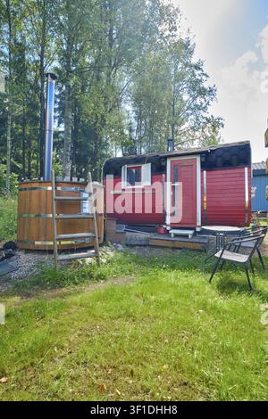 Cabina sauna in legno tra gli alberi con erba verde tutt'intorno. Nelle vicinanze c'è una vasca idromassaggio in legno. Il sole splende, creando un'atmosfera luminosa. Alberi Foto Stock