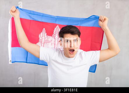 Eccitato, attivo spettatore maschile tiene bandiera nazionale della Cambogia in mani e canti slogan popolare. Foto Stock