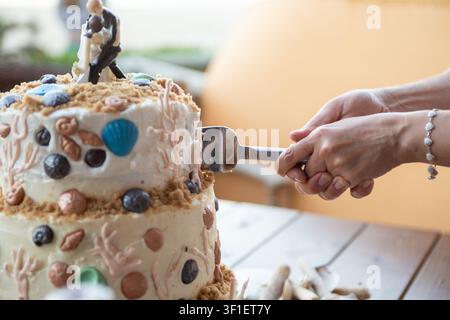 Le mani di una coppia di sposi novelli tagliano una torta nuziale a più piani decorata con conchiglie commestibili e sabbia. Foto Stock