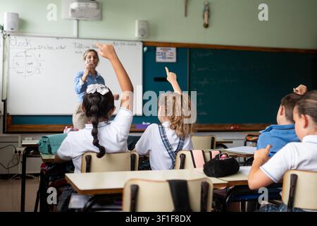 Vista posteriore di bambini delle scuole elementari che alzano le mani durante una lezione in classe con il loro insegnante. Foto Stock