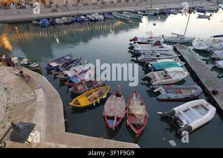 Barche da pesca tradizionali e yacht moderni ormeggiati in un porto sereno con persone che si rilassano lungo il lungomare durante l'ora dorata. Italia, Giovinazzo, 1 Foto Stock