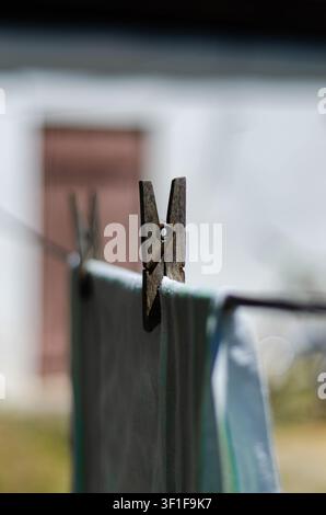Prima piano di una vecchia molletta di legno che tiene un foglio di biancheria bagnata su una corda da bucato contro uno sfondo sfocato di una casa bianca rustica. Stile cottagecore. Foto Stock