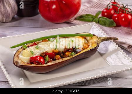 Mezza melanzana al forno ripiena di verdure miste e formaggio fuso, guarnita con cipolla verde. Servita su piatto di ceramica bianca con basilico al pomodoro sullo sfondo Foto Stock