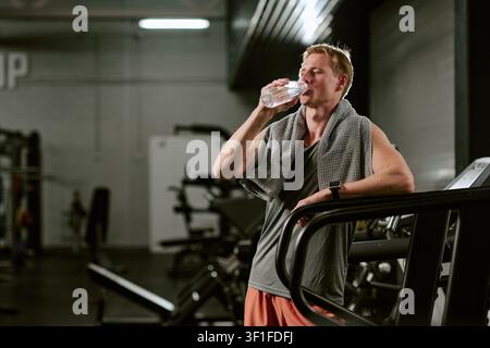 Giovane adulto che beve acqua dopo un allenamento cardio in palestra Foto Stock
