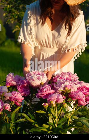Una donna con un vestito bianco e un cappello di paglia tocca delicatamente un grande mazzo di peonie rosa vivaci in un contesto di giardino illuminato dal sole Foto Stock