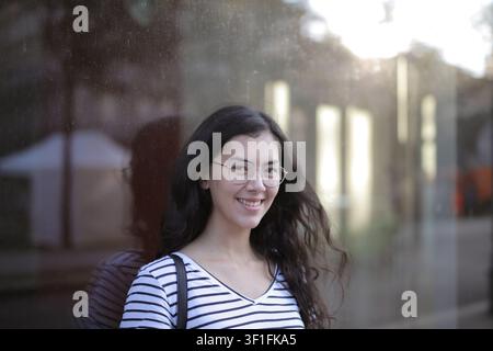 Giovane donna sorridente con gli occhiali in piedi vicino a una finestra Foto Stock
