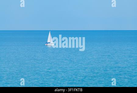 Barca a vela bianca che naviga sul mare blu profondo sotto un cielo blu sereno Foto Stock