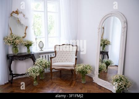 Una stanza luminosa e romantica con una classica poltrona trapuntata, un tavolo console in legno scuro ornato e diverse vasi di vetro pieni di camomilla selvatica Foto Stock
