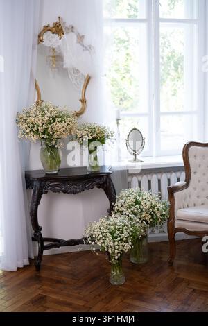 Una stanza luminosa e romantica con una classica poltrona trapuntata, un tavolo console in legno scuro ornato e diverse vasi di vetro pieni di camomilla selvatica Foto Stock