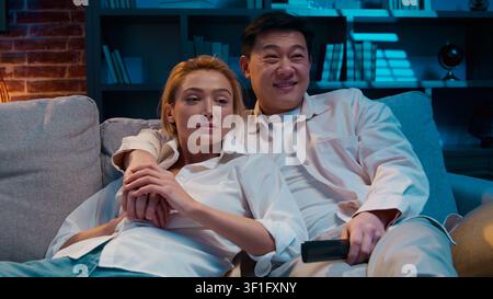 Tranquilla coppia multirazziale che guarda il cinema e la televisione coccolarsi riposando in un comodo soggiorno sul divano la sera a casa di un uomo asiatico eterogeneo Foto Stock