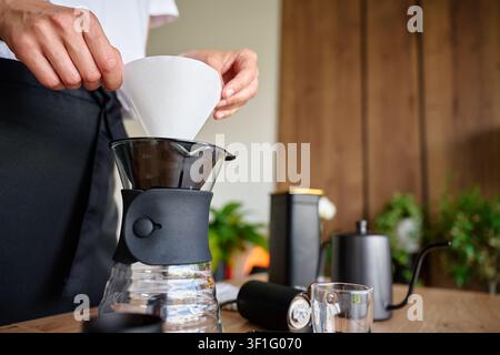 Donna che prepara il caffè filtro a casa, inserendo un filtro di carta nel dripper per il caffè con bollitore e attrezzi per il caffè sul tavolo. Concetto di caffè filtro, preparazione manuale e bevanda mattutina Foto Stock