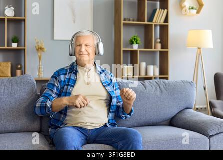 Uomo anziano felice con cuffie sul divano a casa, rilassandosi e godendo della musica Foto Stock