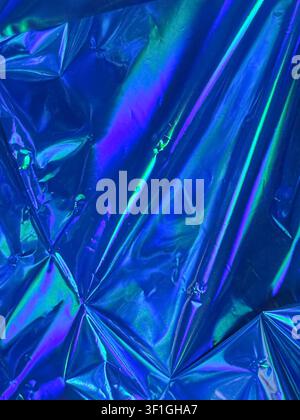 Materiale olografico lucido accartocciato che mostra riflessi iridescenti blu, viola e verdi vivaci. Texture creata dall'interazione della luce su una superficie irregolare Foto Stock