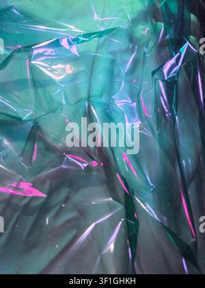 Sfondo astratto con materiale plastico iridescente accartocciato. La superficie traslucida riflette luce magenta e ciano, creando un effetto testurizzato dinamico Foto Stock