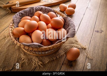 Cesto pieno di uova marroni fresche su un tavolo di legno rustico con una frusta e frammenti di guscio Foto Stock