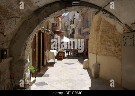 Arco della vecchia città di Bari che conduce in un vicolo lastricato stretto con persone che passeggiano, edifici storici in pietra e biancheria appesa. Italia, Bari, 21 luglio 202 Foto Stock