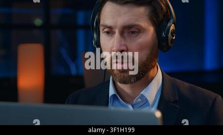 Uomo d'affari caucasico concentrato con cuffie, operatore maschile che parla al telefono in un call center alla sera, uomo d'affari di mezza età che lavora in un call center Foto Stock