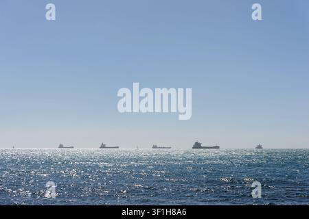 Flotta di navi da carico e imbarcazioni ancorate in mare aperto sulla linea dell'orizzonte sotto un cielo blu terso con riflessi del sole sull'acqua Foto Stock