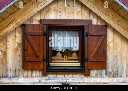 Finestra di capanna in legno rustica con persiane aperte. Vista frontale di una finestra di capanna in legno rustica con persiane marroni aperte, tende di pizzo e legno a vista Foto Stock