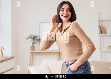 Giovane donna sorridente con gli occhi chiusi che ascolta attentamente con la mano a coppa all'orecchio in un interno luminoso, indossa una maglietta casual e jeans Foto Stock