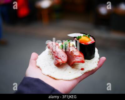 Una mano tiene un pezzo di nigiri di manzo Hida wagyu su un cracker di riso acquistato da un banco di un mercato di strada giapponese, con altri alimenti e lo sfondo sfocato del mercato visibile. Foto Stock