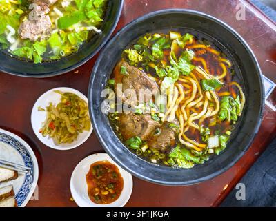 Una vista dall'alto di una zuppa di noodles con manzo brasato in una pentola di argilla nera con noodles spessi, manzo brasato e verdure a foglia in brodo di soia scuro, accompagnata da verdure sottaceto e salsa al peperoncino in un ristorante a Kaohsiung, Taiwan. Foto Stock
