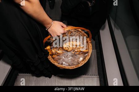 Vista dall'alto di una donna che tiene un cesto di vimini pieno di biscotti decorati per Halloween avvolti in sacchetti regalo trasparenti mentre sale su una scala mobile. Concetto di prelibatezze stagionali, shopping festivo e celebrazione delle festività. Foto Stock
