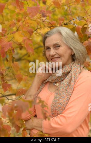Donna sorridente che sistema la sciarpa tra il fogliame autunnale con un'atmosfera felice e rilassata in un primo piano, vista leggermente inclinata verso l'alto della moda stagionale Foto Stock