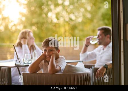 Ragazzo sorridente che si rilassa sul balcone di casa con i genitori che godono di bevande serali e della vista sulla terrazza estiva Foto Stock