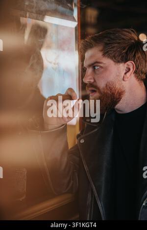 Uomo barbuto che fuma e guarda attraverso una finestra in un'atmosfera suggestiva da bar Foto Stock