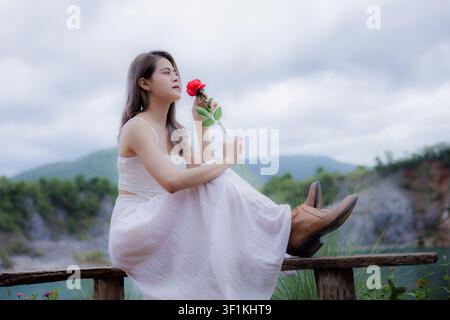 Una giovane donna graziosa in un vestito estivo bianco posa all'aperto tenendo una rosa rossa con un paesaggio naturale sfocato in sottofondo. Foto Stock