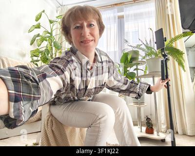 Donna seduta comodamente nel suo salotto che scatta un selfie circondata da piante d'appartamento, catturando un momento della sua vita quotidiana. Foto Stock