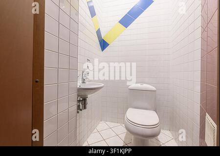 piccolo bagno con piastrelle bianche, un lavandino e una toilette. La parete ha un motivo geometrico blu e giallo Foto Stock