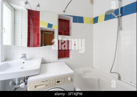 Bagno luminoso con piastrelle quadrate bianche. Le caratteristiche includono una vasca da bagno, un lavandino, una lavatrice, uno specchio con accenti rossi e dettagli piastrellati blu/gialli Foto Stock