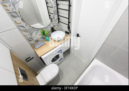 Bagno compatto con impianti moderni come uno specchio rotondo, un lavandino sospeso, una lavatrice e una parete geometrica con piastrelle decorative Foto Stock