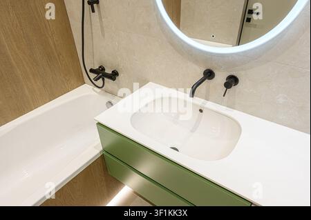 Bagno luminoso con lavandino bianco, armadi verdi, rubinetteria nera, vasca da bagno, pareti piastrellate e specchio circolare retroilluminato Foto Stock