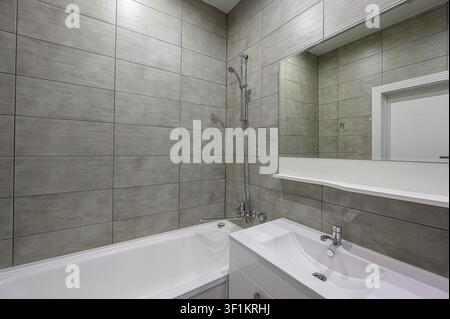 bagno moderno con pareti piastrellate grigie, una vasca bianca con doccia e un lavandino bianco elegante con un grande specchio sopra Foto Stock