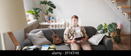 Ritratto di un giovane uomo in salotto, seduto con un tablet in mano, ha la valigia imballata e due biglietti aerei, si prepara ad andare all'estero, andando avanti Foto Stock