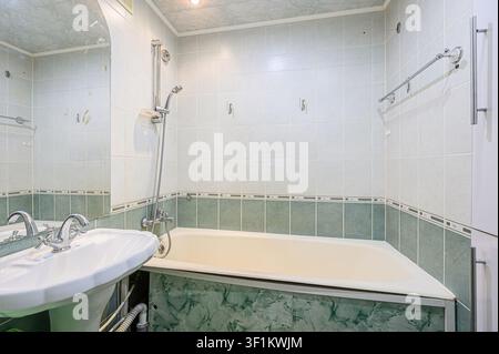 Bagno con piastrelle chiaro, un lavandino bianco, una vasca da bagno e rubinetteria argentata Foto Stock