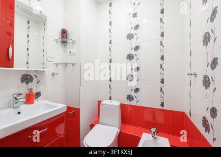 bagno luminoso con pareti piastrellate bianche con accenti floreali, un lavabo rosso, WC e bidet. Ha un design moderno e pulito Foto Stock
