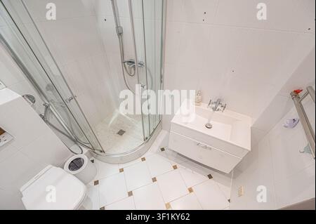 piccolo bagno con piastrelle bianche dotato di cabina doccia in vetro, toilette e lavandino moderno bianco. Le piastrelle del pavimento sono bianche con decorazione ac Foto Stock
