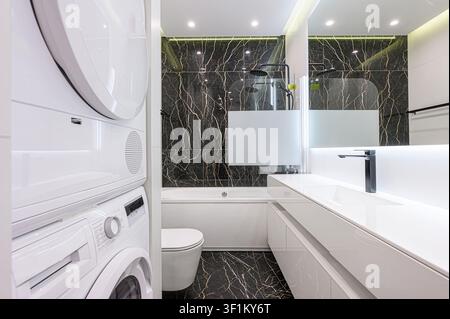 Bagno elegante e moderno con pareti/pavimenti a motivi di marmo nero, contrastati da elementi bianchi, tra cui una lavatrice/asciugatrice impilata, un WC e un vani Foto Stock