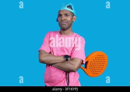 Giovane uomo in piedi con le braccia incrociate, che tiene una racchetta da padel, mostrando un'emozione scettica Foto Stock