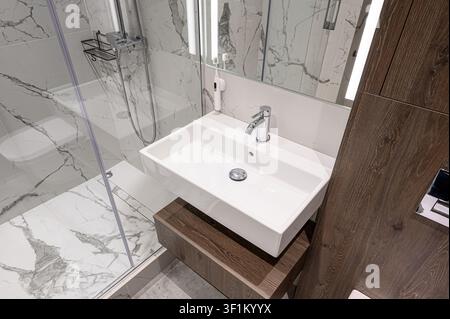 Bagno moderno con lavandino rettangolare bianco, rubinetto cromato, pareti e pavimento a motivi di marmo e un armadio di legno. L'area della doccia è visibile su t Foto Stock
