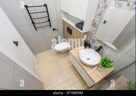 bagno moderno con un lavandino bianco su un mobile tono legno, pareti piastrellate grigie, un gabinetto bianco, un riscaldatore per asciugamani nero e un accento decorativo w Foto Stock