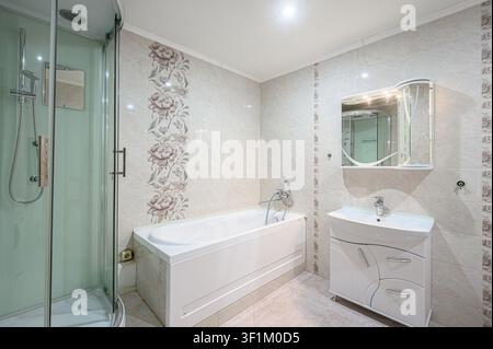 Bagno luminoso con vasca, doccia in vetro, lavandino con lavabo e pareti piastrellate nei toni del beige con accenti floreali Foto Stock