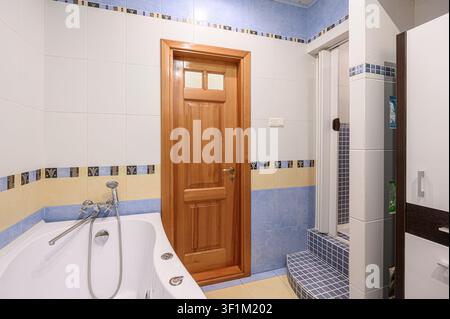 bagno ben illuminato con una vasca bianca, pareti piastrellate con accenti blu e gialli, una porta in legno e una doccia walk-in con piastrelle a mosaico blu Foto Stock
