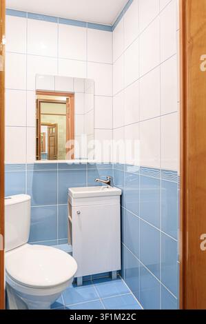 Viene mostrato un piccolo bagno con piastrelle blu e bianche. Sono visibili un gabinetto e un mobile per il lavandino. Uno specchio riflette una porta che conduce ad un'altra stanza Foto Stock